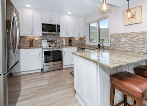 A modern kitchen with white cabinetry, stainless steel appliances, a large island with bar stools, and a view of the outdoors through a window.