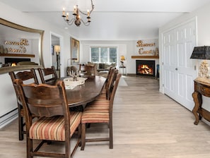 A dining room with a large wooden table, chairs, a chandelier, a fireplace, and a door.