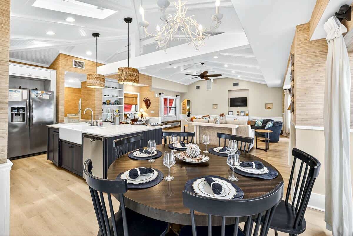 A modern kitchen with a central island, dining area with round table, and a living room with a sofa and TV.