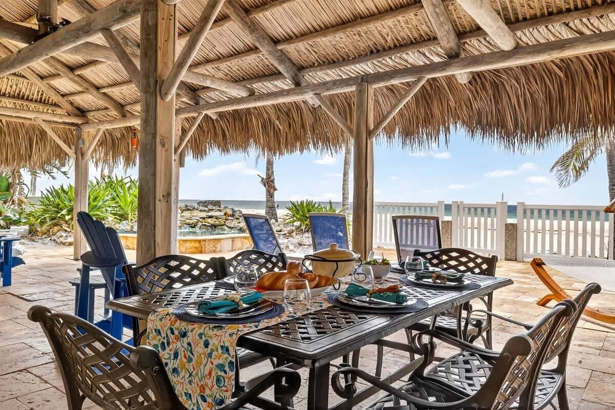 An outdoor dining area with a thatched roof, tables set for two, and chairs.