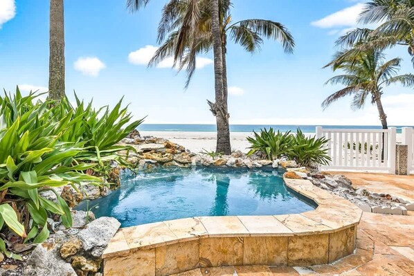Ein Pool mit Steinrand, umgeben von tropischen Pflanzen und Palmen, mit Strand und Meer im Hintergrund.