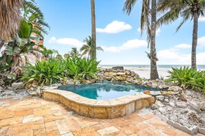 Ein Pool, umgeben von Felsen und Pflanzen, mit Strand und Palmen im Hintergrund.