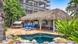 A pool area with lounge chairs, a thatched-roof structure, and a building under construction in the background. - Gulf Front Dreams Beachfront Home in Clearwater (Clearwater Beach)