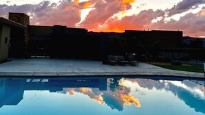Ein Swimmingpool spiegelt einen Sonnenuntergangshimmel mit Wolken wider, umgeben von Gebäuden und einer gepflasterten Fläche.