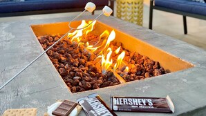Ein Lagerfeuer mit Marshmallows und Schokoriegeln, umgeben von Sitzgelegenheiten mit gemusterten Kissen.