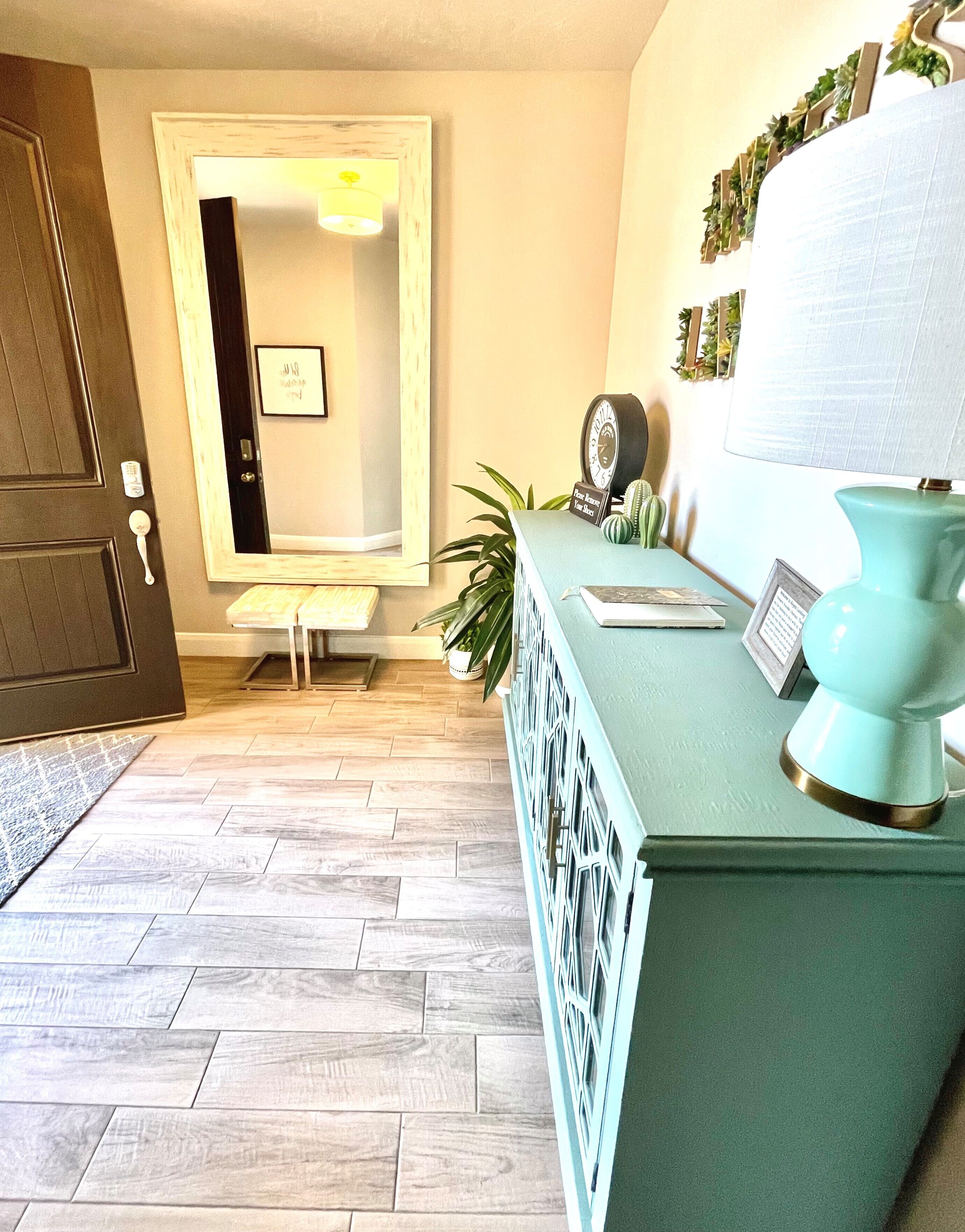 A room with a wooden door, a mirror, a teal dresser with a clock, a plant, and a framed picture.