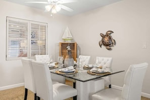 A dining area with a glass table, white chairs, a ceiling fan, and a wall-mounted turtle sculpture.