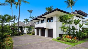 A modern two-story house with a white exterior and dark roof, surrounded by lush tropical vegetation.