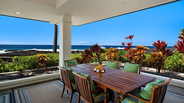 A wooden table with green cushions, surrounded by tropical plants, overlooking a beach with the ocean and rocky shoreline.