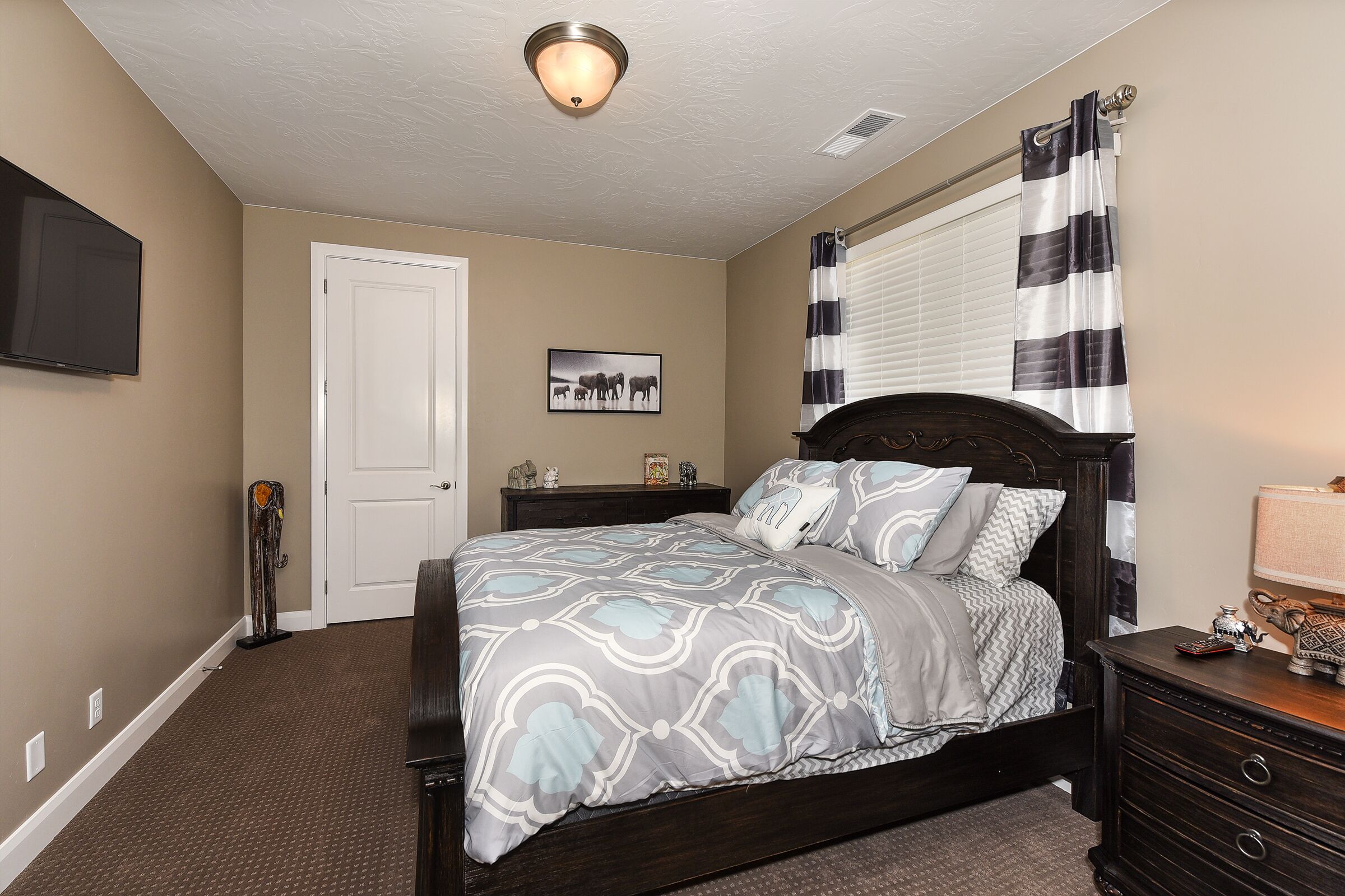 A bedroom with a dark wooden bed, a flat-screen TV mounted on the wall, a door, a window with black and white curtains, and a small table with a lamp.