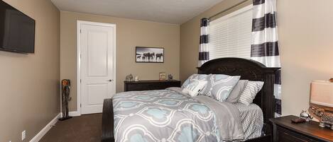 A bedroom with a dark wooden bed, a flat-screen TV mounted on the wall, a door, a window with black and white curtains, and a small table with a lamp.