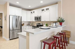 A modern kitchen with white cabinets, stainless steel appliances, a central island with stools, and a potted plant.