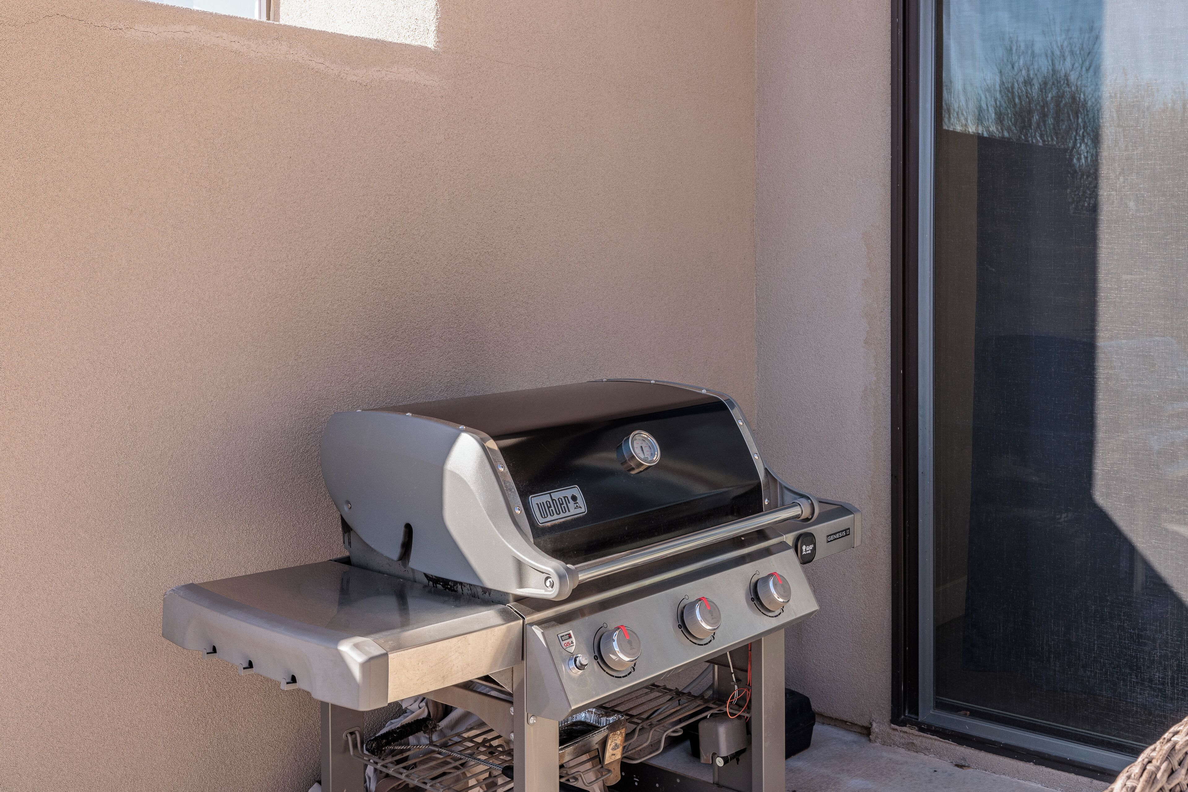 A portable gas grill with four burners, a digital display, and a side shelf, positioned against a wall next to a sliding glass door.
