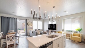 Une cuisine moderne avec un îlot central, un coin repas avec des chaises blanches, un salon avec un canapé et une télévision, et un fauteuil jaune.