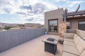 Un patio en la azotea con chimenea de piedra, mobiliario exterior y vistas de los edificios y el paisaje circundante.