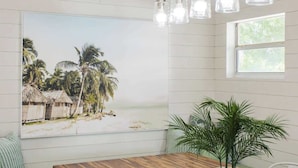A room with a wooden ceiling, a wooden dining table with a plant centerpiece, a large beach-themed painting, and a window with natural light.