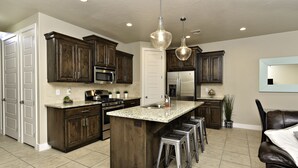 A spacious kitchen with dark wooden cabinets, a large island with stools, stainless steel appliances, and pendant lighting. - Sweeping Red Rock Views - 4BR close to Pool & Hot Tub (St. George)