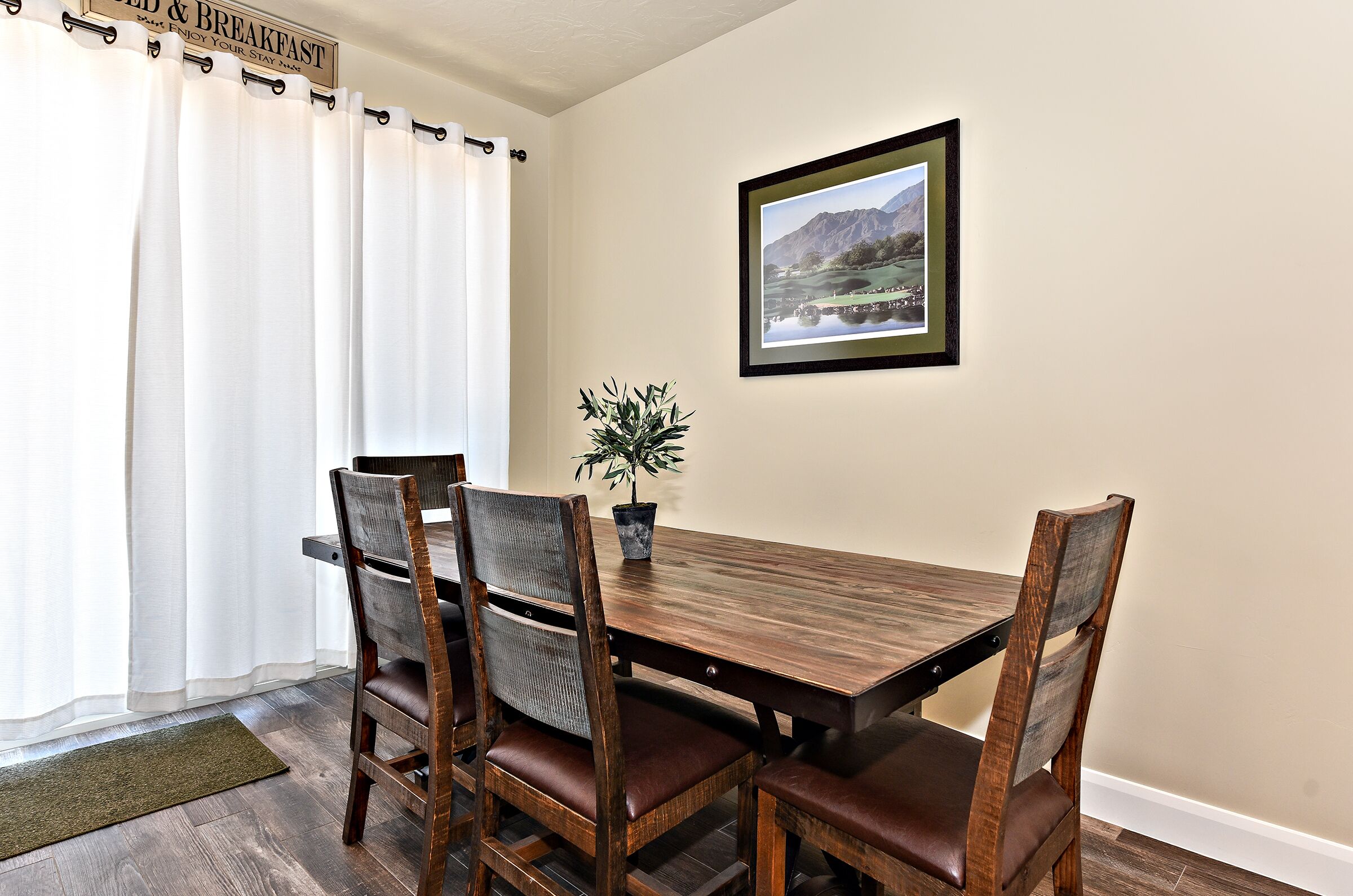 A dining area with a wooden table and four chairs, a potted plant, and a framed picture on the wall.