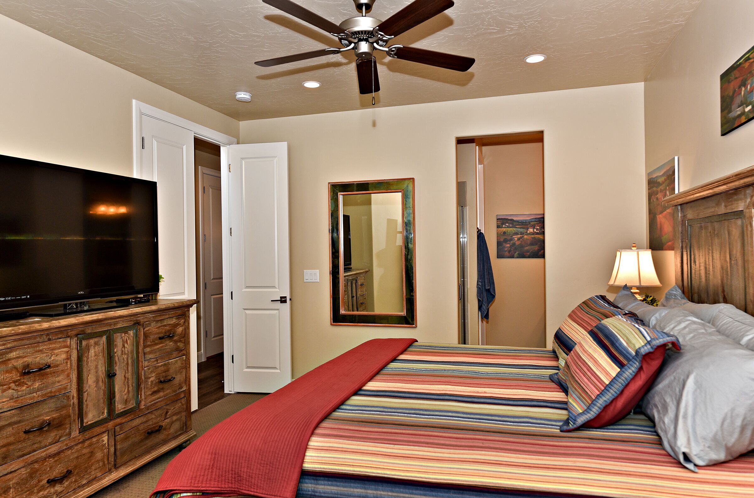 A bedroom with a TV on a wooden cabinet, a bed with striped bedding, a ceiling fan, and a mirror on the wall.