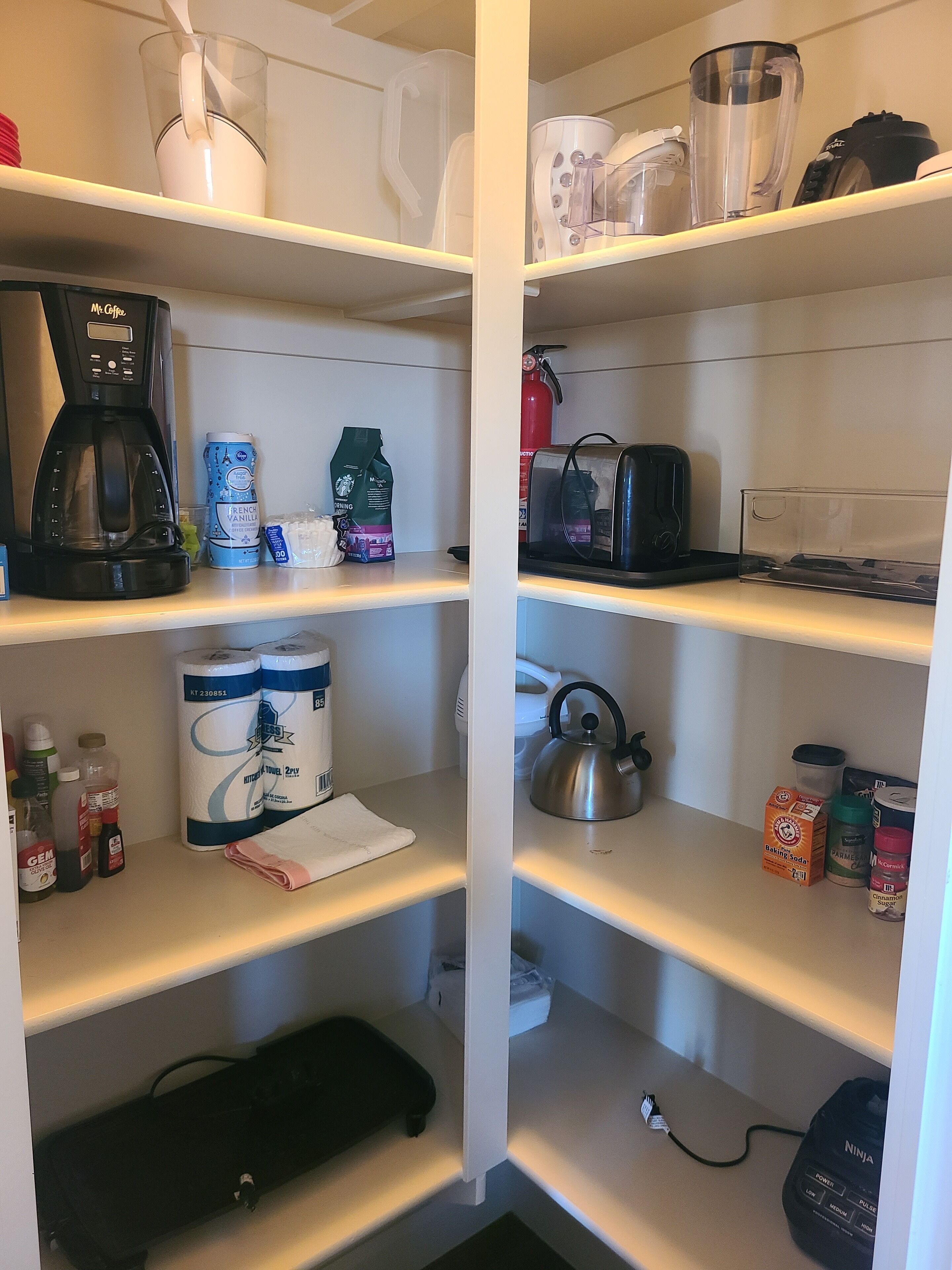A kitchen with shelves stocked with coffee maker, coffee pods, paper towels, a kettle, and various kitchen utensils.