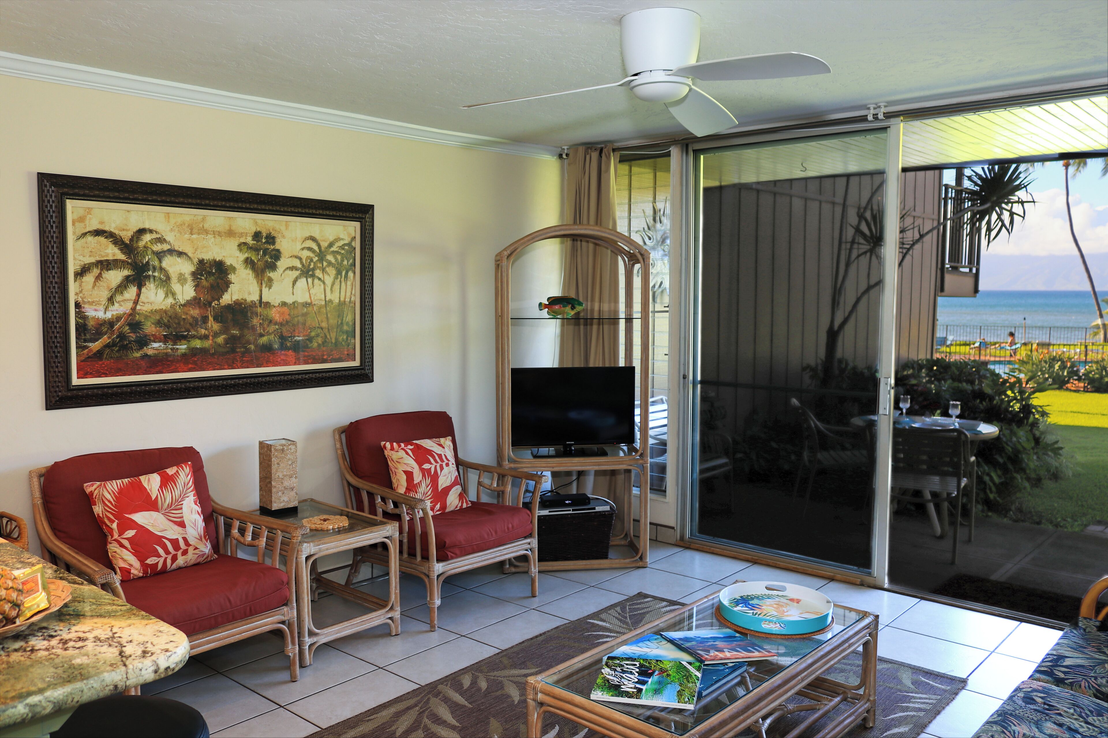A living room with a television, red cushioned chairs, a glass coffee table, and a large painting of a tropical scene.