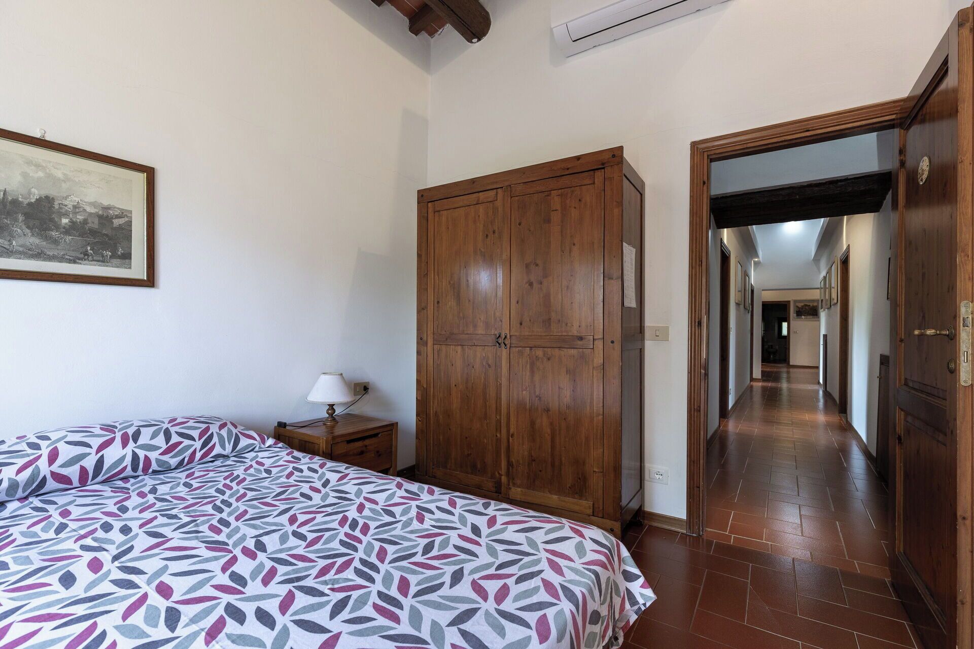 A bedroom with a bed, a wooden wardrobe, a nightstand with a lamp, and a hallway leading to another room.