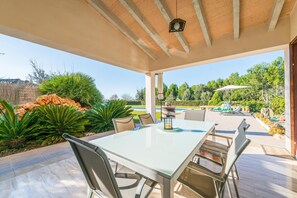 Alpendre coberto com mesa e cadeiras, rodeado por vegetação exuberante e vista para uma piscina.