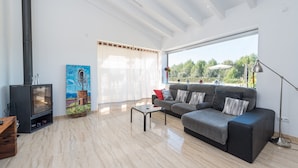 A modern living room with a sofa, a coffee table, a fireplace, and a large window offering a view of greenery. - ES SERRAL - Villa with private pool in sa Pobla. Free WiFi (Sa Pobla)