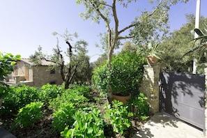 A stone house with a tiled roof, surrounded by lush greenery and a metal gate.