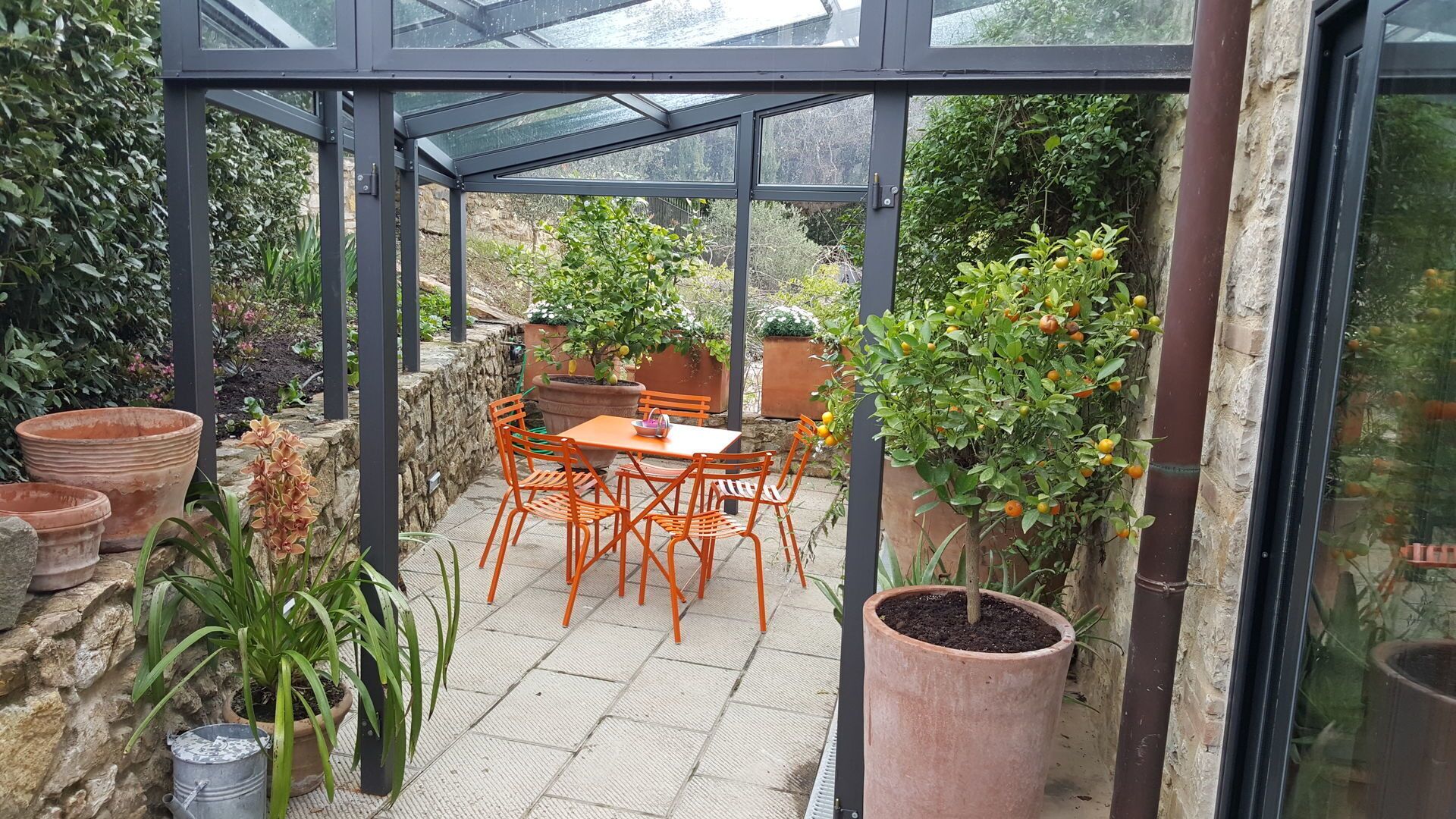 Une véranda avec un toit en verre, un mur en pierre et une table avec des chaises. On y trouve des plantes en pot et un grand pot en terre cuite.