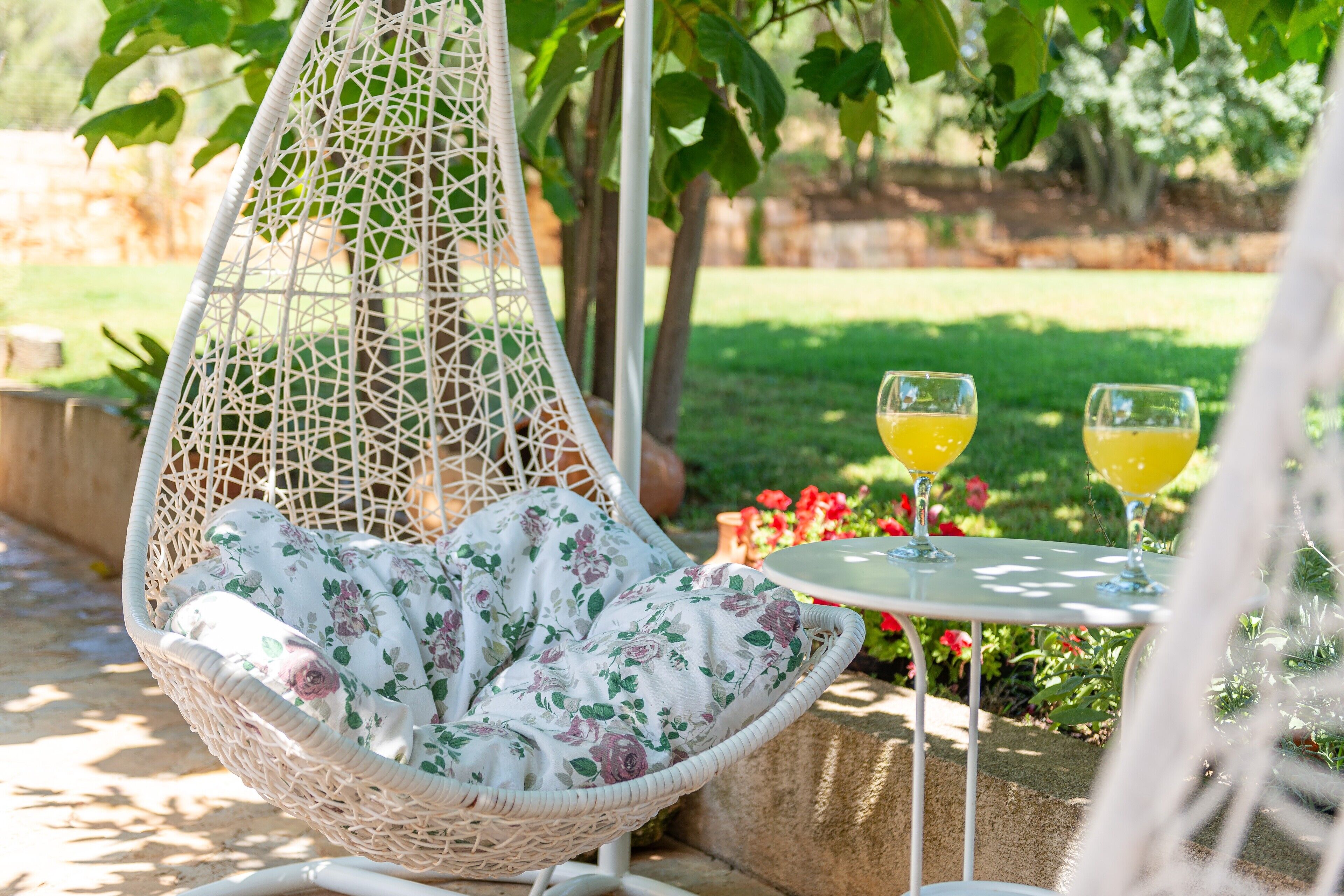 A hanging chair with a floral cushion, a table with two glasses of a yellow beverage, and a garden with red flowers and green grass.
