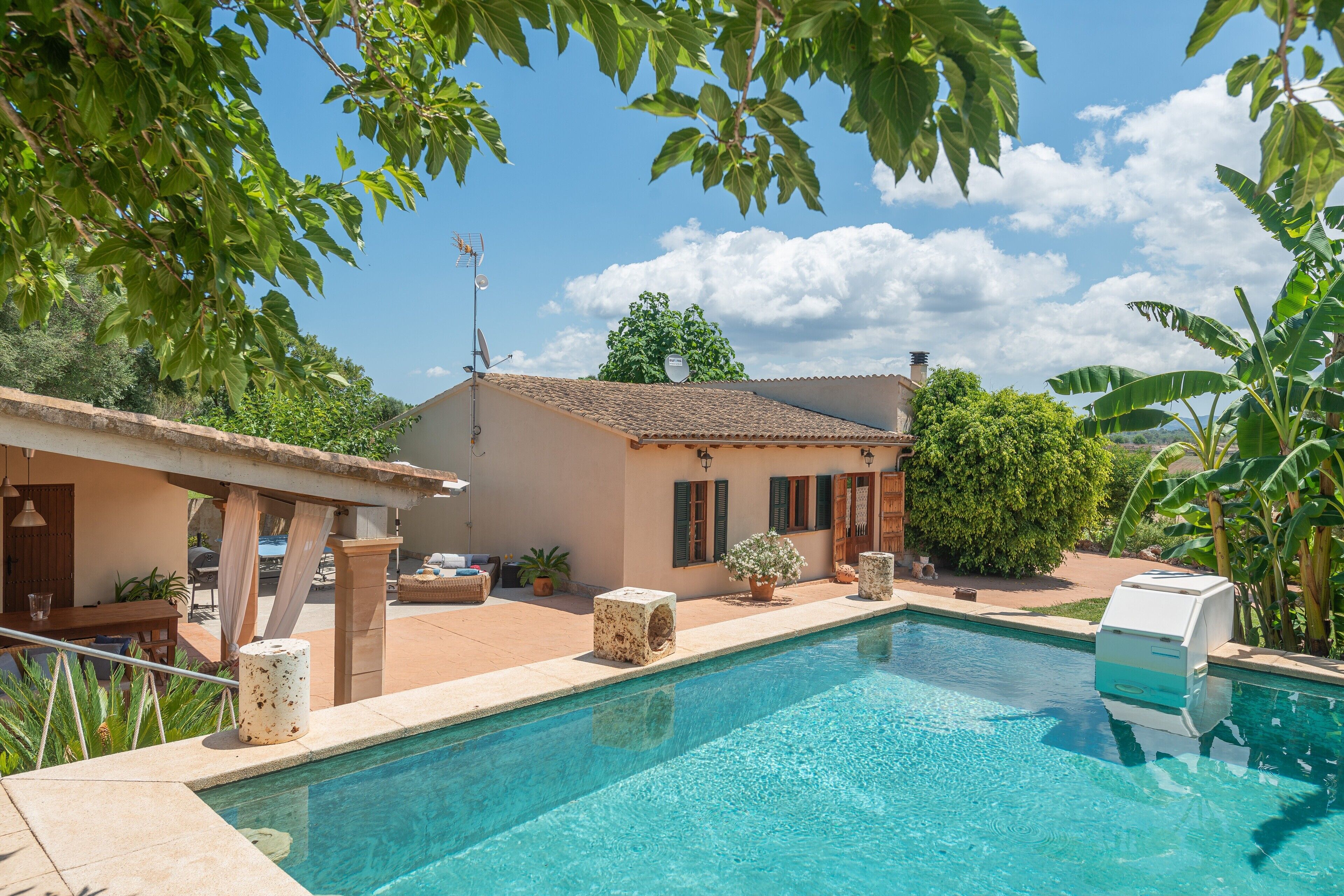 A swimming pool with a stone structure, a house with a tiled roof, and surrounding greenery.