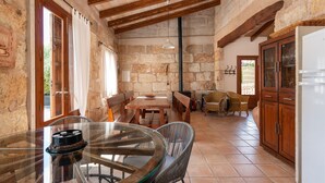 A rustic stone interior with a glass table, wooden benches, wicker chairs, and a kitchen area with wooden cabinets and a refrigerator. - ALCUDIOLA - Villa with private pool in Santa Margarita. Free WiFi (Santa Margalida)