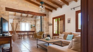 A spacious living area with a sofa, coffee table, and a television. There is a dining table with chairs and a stone wall in the background. - ALCUDIOLA - Villa with private pool in Santa Margarita. Free WiFi (Santa Margalida)