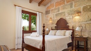 A bedroom with a wooden four-poster bed, white linens, a stone wall, a window with curtains, and a nightstand with a lamp. - ALCUDIOLA - Villa with private pool in Santa Margarita. Free WiFi (Santa Margalida)