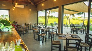 Restaurante com mesas e cadeiras dispostas para refeições, grandes janelas com vista para um campo de golfe e luminárias pendentes.