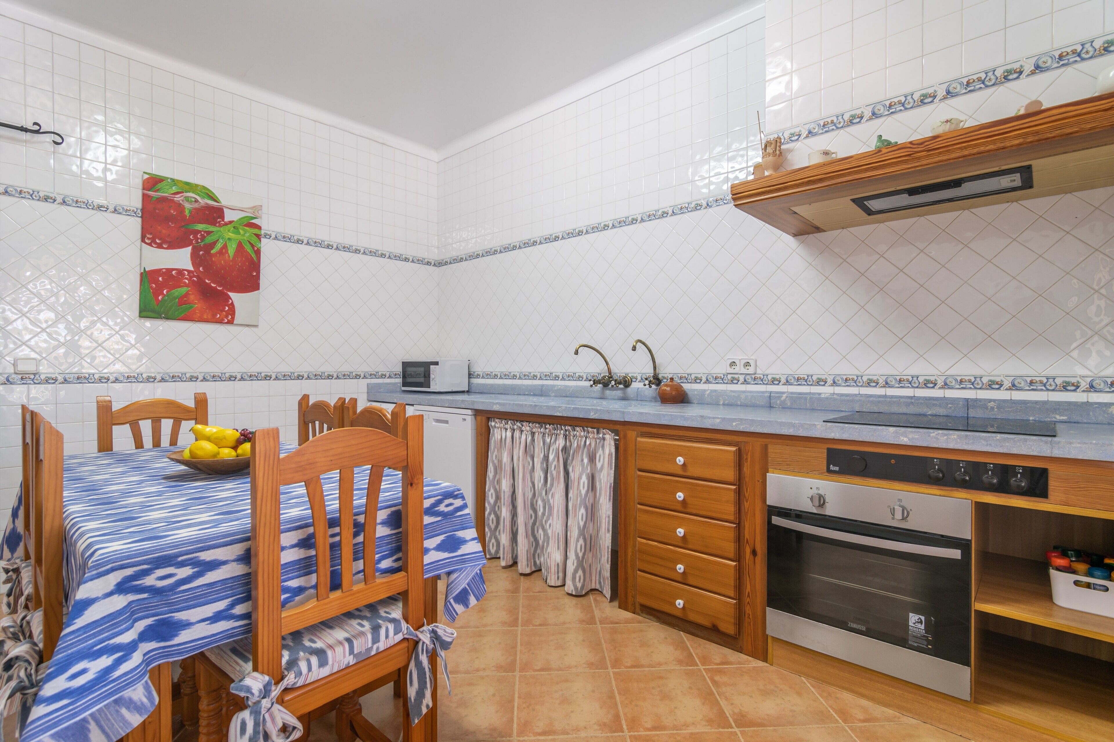 A kitchen with white tiled walls, a wooden dining table with a blue and white tablecloth, a built-in oven, and a microwave.