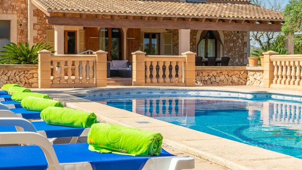 A pool area with lounge chairs and towels, a stone building with a tiled roof, and a clear blue pool.