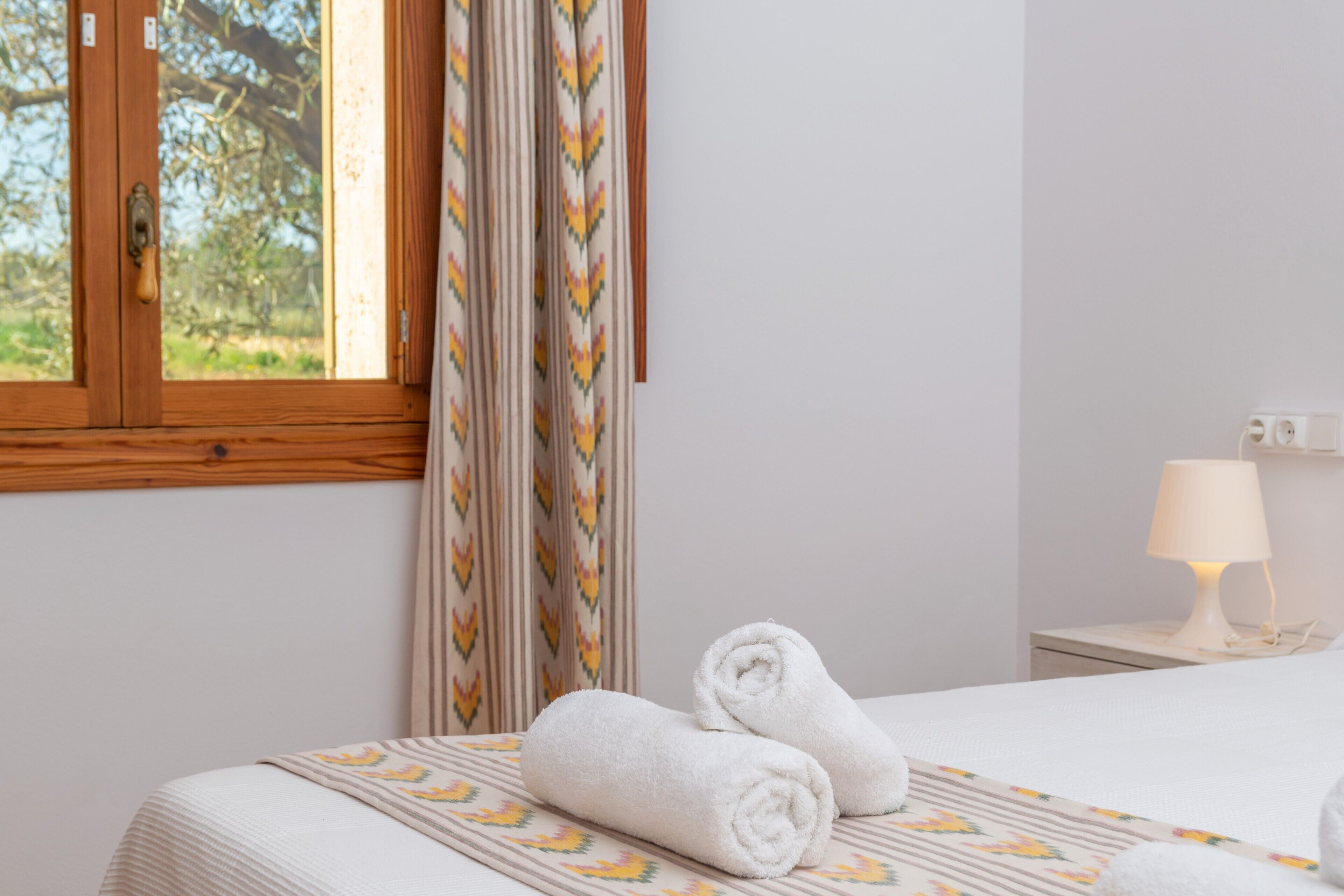 A hotel room with a bed, a window with curtains, a bedside lamp, and a view of trees outside.