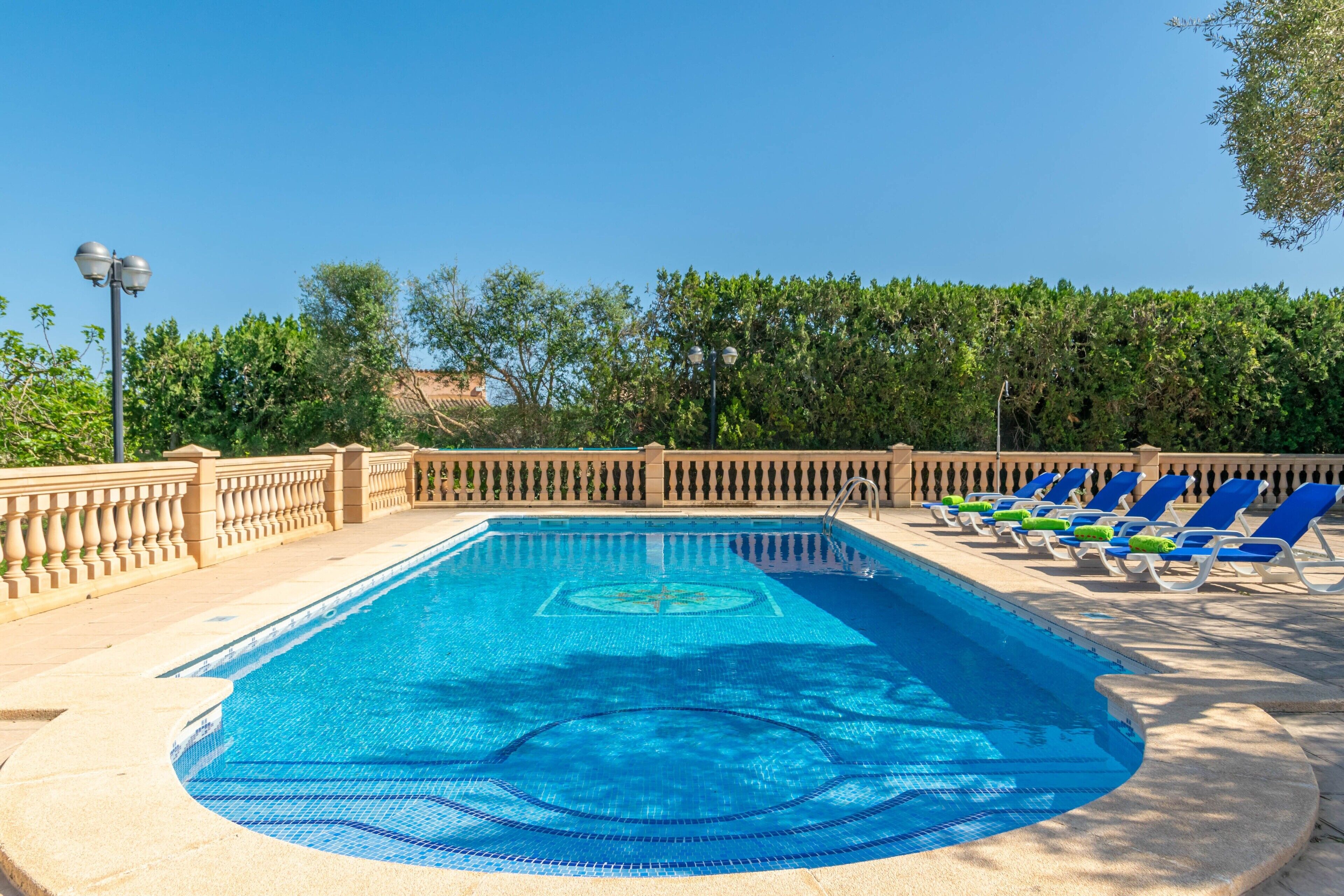 A rectangular swimming pool with a tiled pattern, surrounded by a stone deck and a low stone wall. There are several blue lounge chairs with cushions along the poolside.
