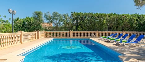 A rectangular swimming pool with a tiled pattern, surrounded by a stone deck and a low stone wall. There are several blue lounge chairs with cushions along the poolside.