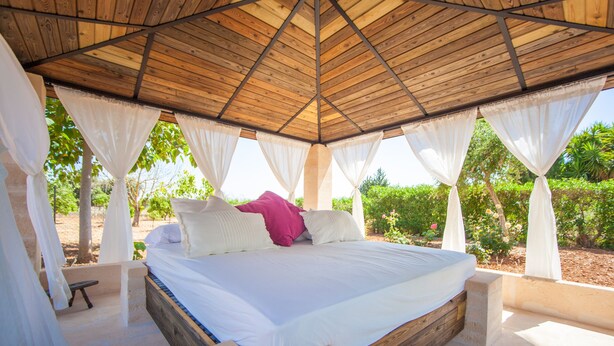 A bed with white bedding and a pink pillow, under a wooden ceiling with white curtains.