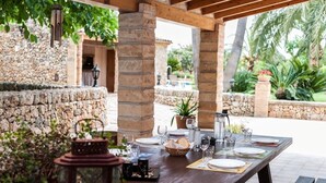 An outdoor dining area with stone walls, wooden pillars, and a table set for two with plates, glasses, and a basket of bread.