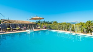 Ein Schwimmbecken mit Liegestühlen und Sonnenschirmen, umgeben von einem Zaun und Bäumen, mit Bergen im Hintergrund.