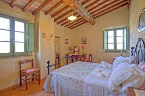 Une chambre avec un plafond en bois, un lit recouvert d’une couvre-lit à motifs floraux, une chaise, un bureau avec un miroir et deux fenêtres dotées de volets verts.