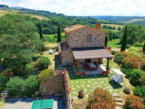 Une maison en pierre dotée d’un patio, entourée d’une végétation luxuriante et avec des collines en arrière-plan.