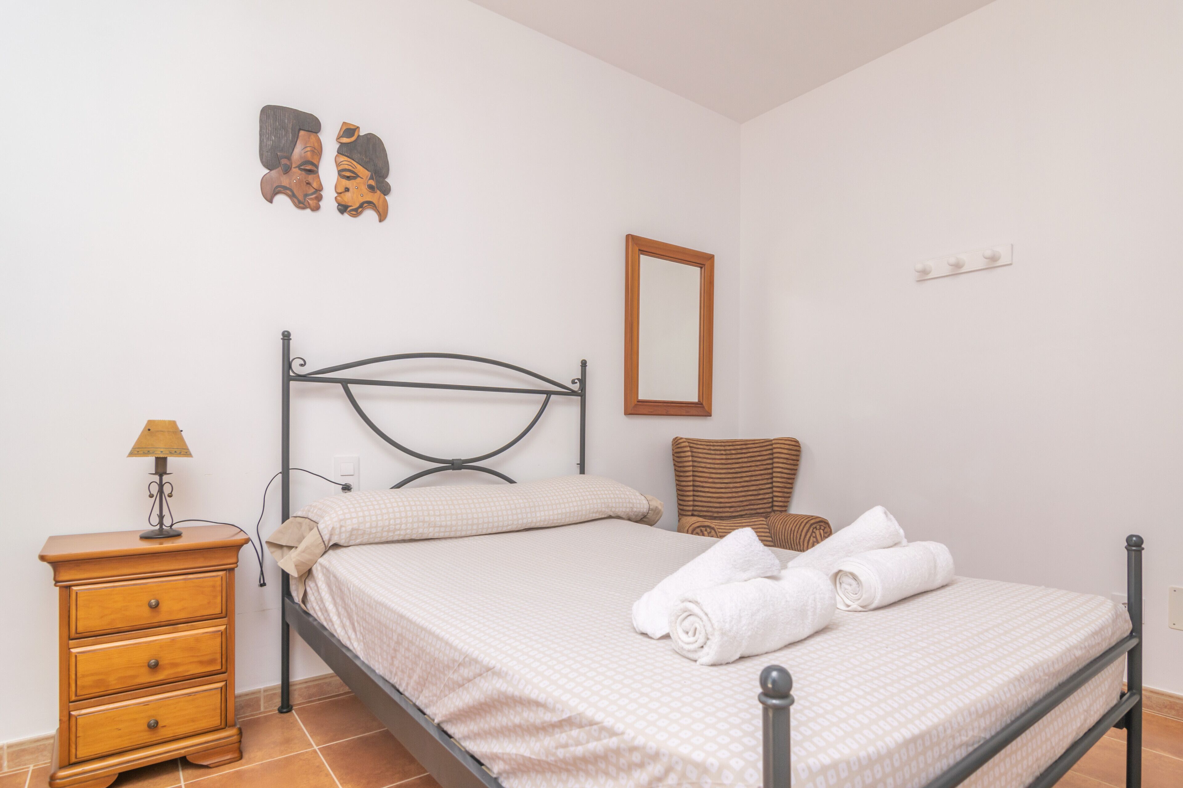 A neatly made bed with a headboard, a wooden nightstand with a lamp, a wicker chair, and two wall-mounted decorations.
