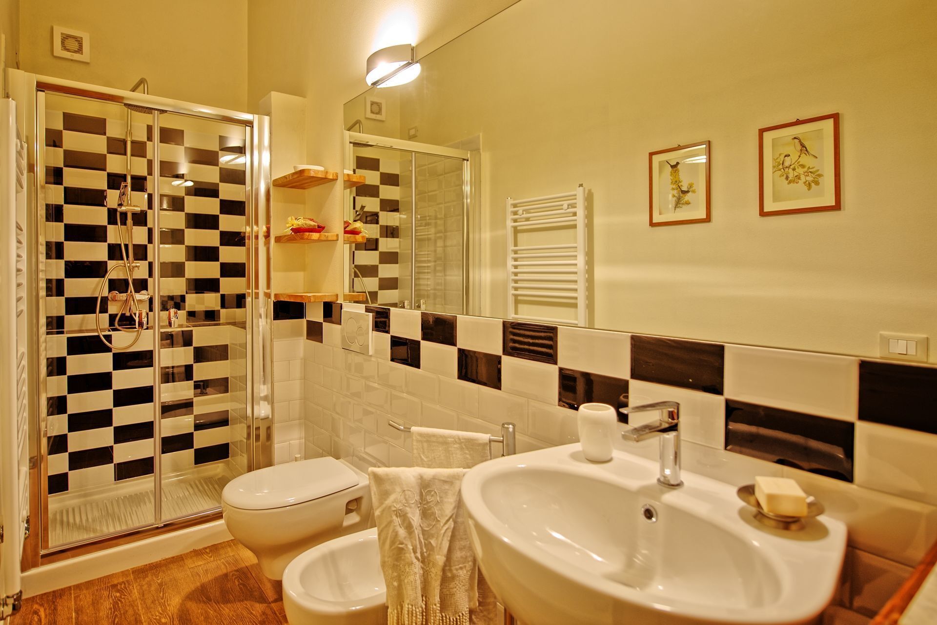 Une salle de bain avec un mur recouvert de carreaux à motifs, une douche en verre, des toilettes, un lavabo avec un miroir et deux tableaux encadrés accrochés au mur.