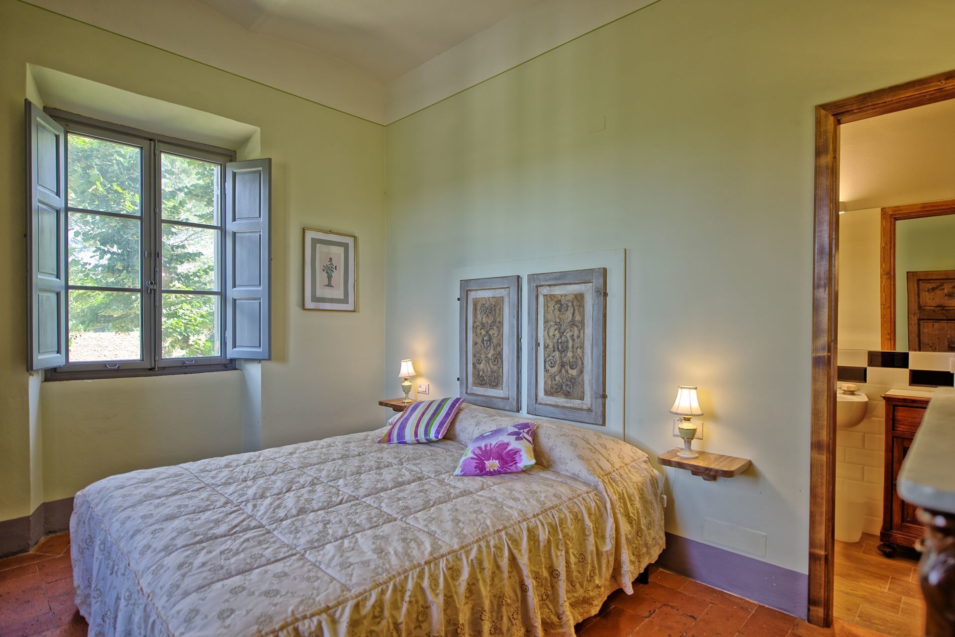 A bedroom with a bed, two bedside lamps, a window with open shutters, and a door leading to another room.