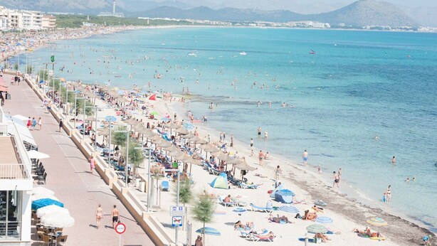 Ein überfüllter Strand mit badenden und sonnenden Personen, ein klares blaues Meer und Berge im Hintergrund.